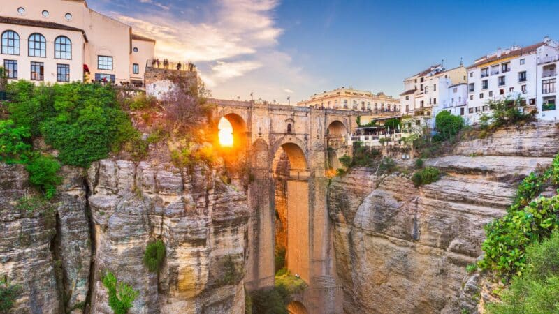 an archway cut into the cliffs surrounded by houses with the sunset peeping through the archway